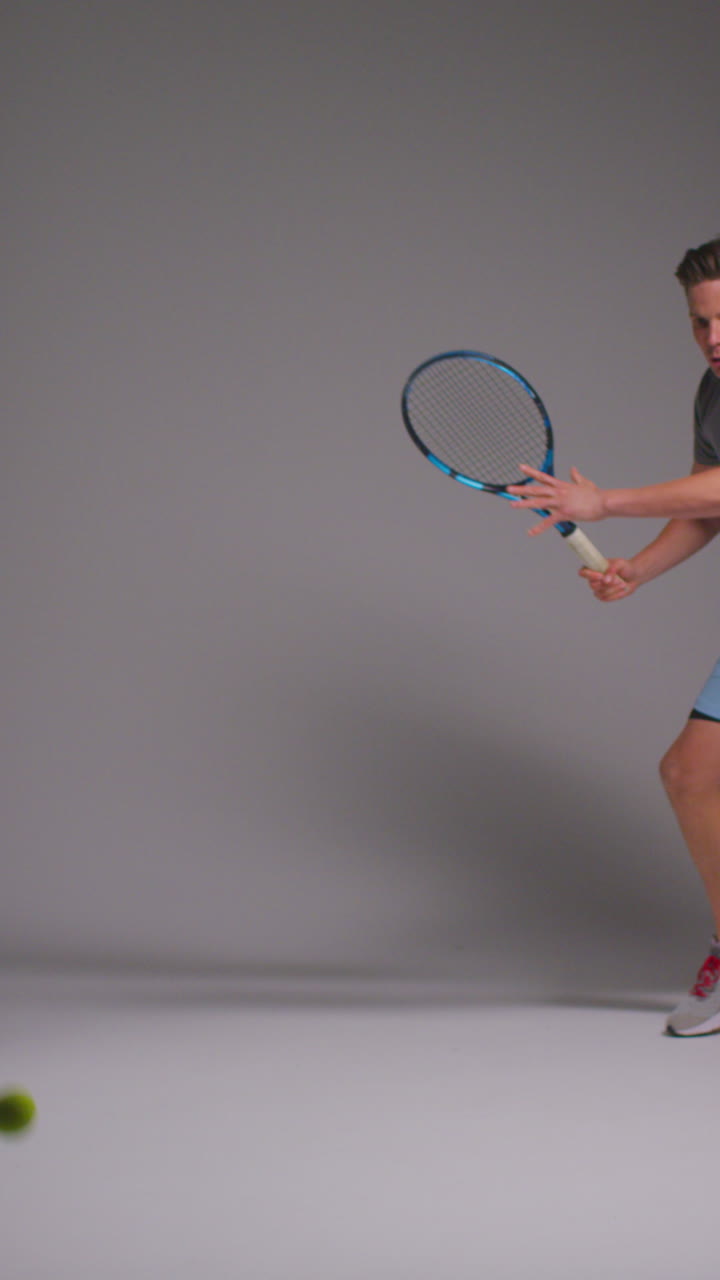 Vertical Video Studio Shot Of Male Tennis Player Returning Shot In Match Hitting Ball With Racket Against Grey Background