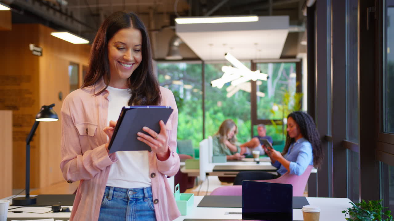 Empresaria de pie junto al escritorio en una oficina moderna de planta abierta usando una tableta digital