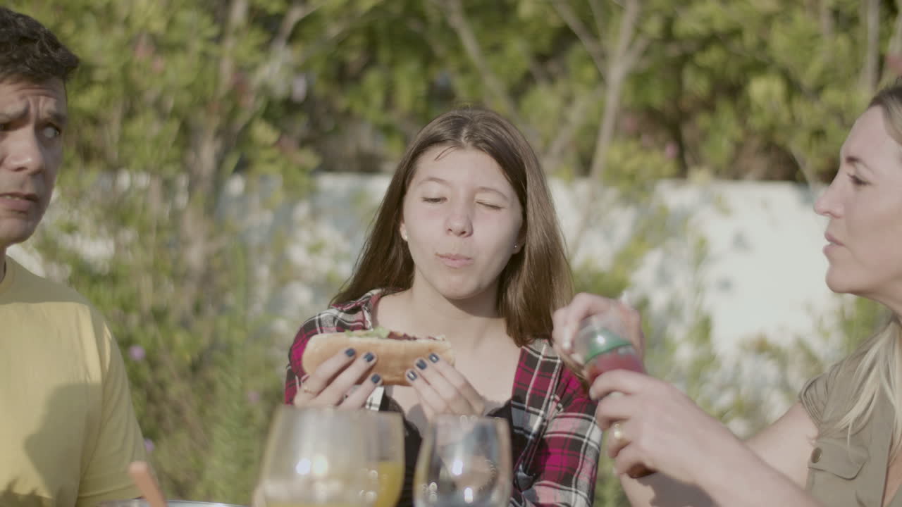 웃고 있는 십대 딸 이 가족 과 함께 점심 식사 할 때 핫도그 를 물고 있다