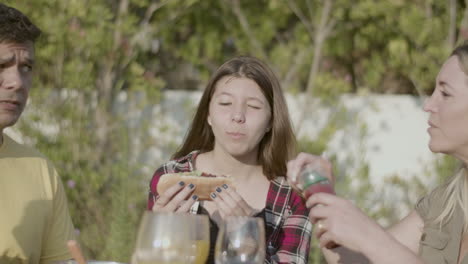 Smiling-teenage-daughter-biting-hotdog-at-lunch-with-family