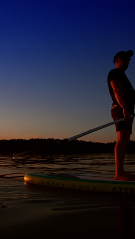 unknown man in cap approaches on the paddle board. sportsman oaring standing on the sup board on the river at sunset. low angle view. vertical video.