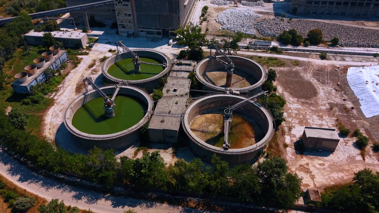 Cuencas industriales circulares con mecanismos centrales. Cuatro tanques de hormigón se alzan junto a edificios de fábrica, carreteras, árboles y grandes pilas de materiales almacenados