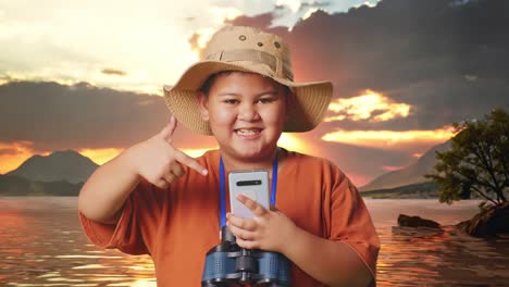 asian boy with a hat and binoculars smiling and pointing to smartphone at a lake. boy researcher examines something, travel tourism adventure concept, close up