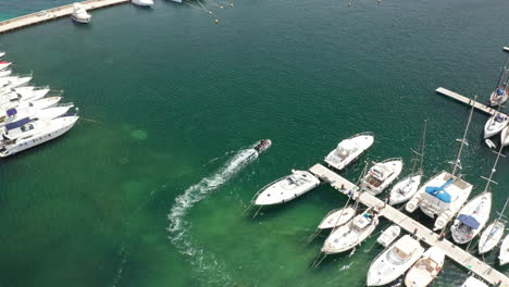 Boats-docked-at-a-marina-with-a-small-motorboat-leaving-the-harbor,-aerial-view