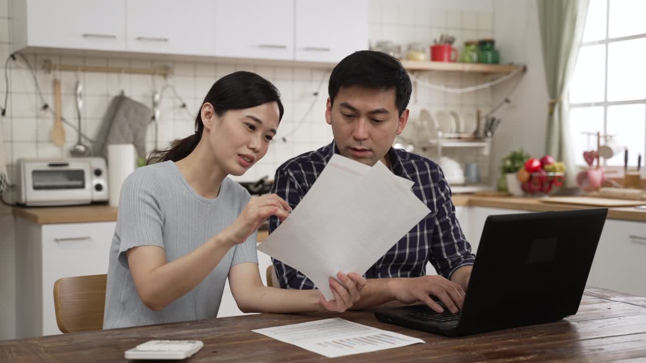 Asian married couple discussing bill for the tax season in the dining room at home. they pointing at computer and looking at bills