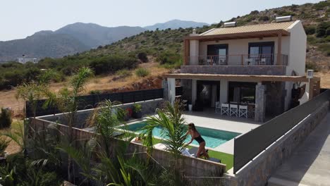 mujer delgada de cabello oscuro camina a una piscina en traje de baño y se acuesta en una cama de sol - se ve una villa en grecia creta y palmeras en primer plano