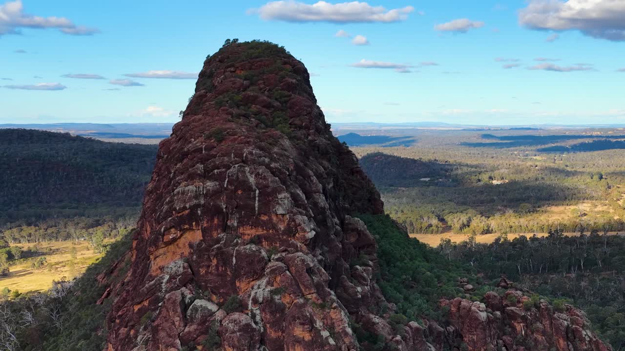 Drone glides over volcanic outcrop, revealing vast Australian landscape under bright daylight and blue sky