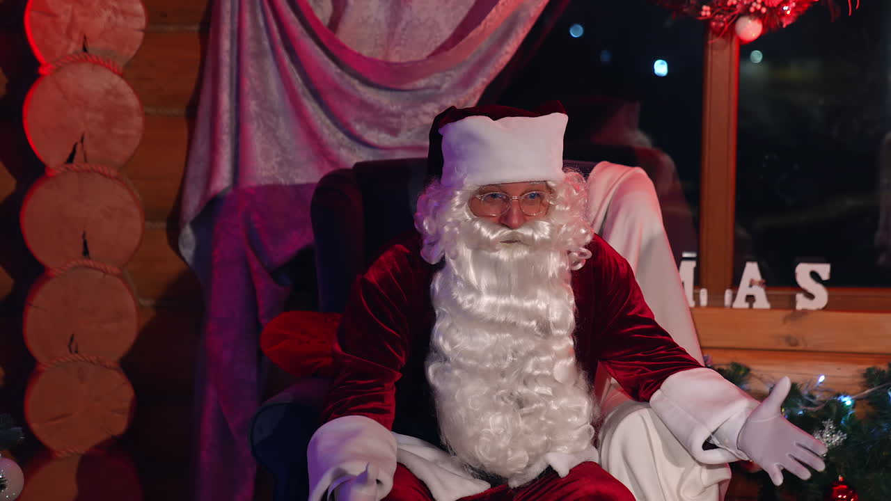 Portrait of Santa Claus in armchair. Real Santa with long white beard and glasses wearing red costume sitting in Christmas decorated room.
