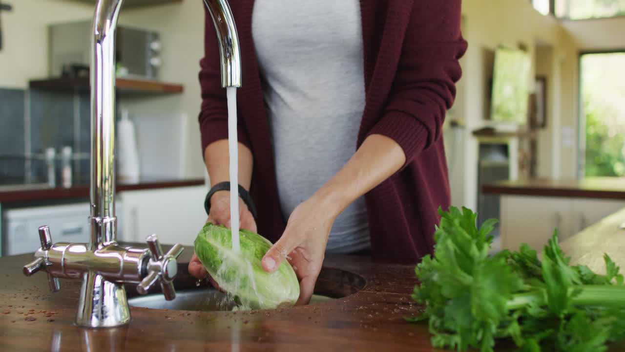 sección media de una mujer caucásica embarazada lavando verduras en la cocina