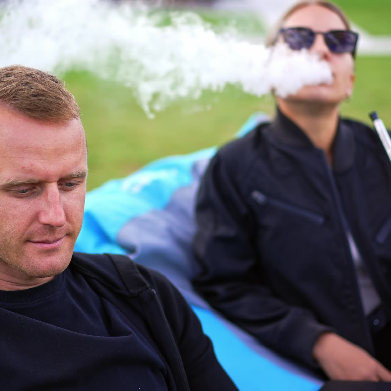 Mid-aged young couple in bean bag chairs resting outdoors. Male is focused on work on laptop and lady is smoking hookah. Summer time rest outdoors