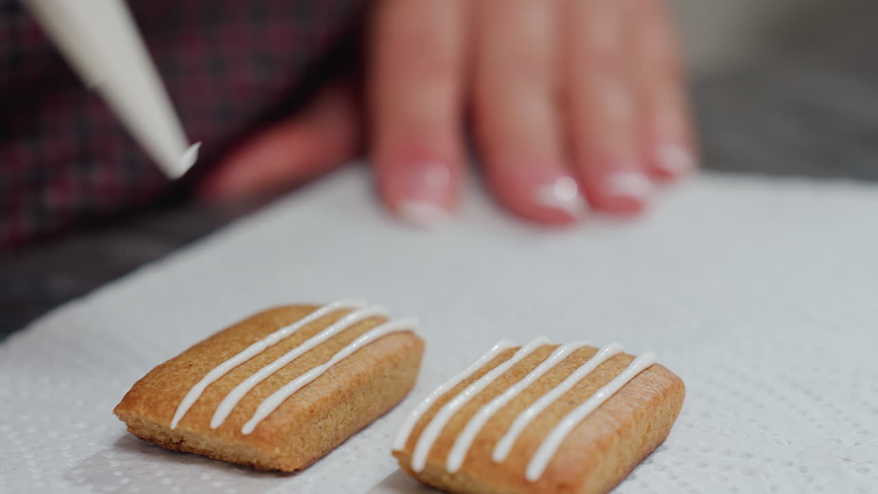 Close-up of baker decorating cookie with white icing lines on tissue paper, neatly manicured nails hold the piping bag for detailed design