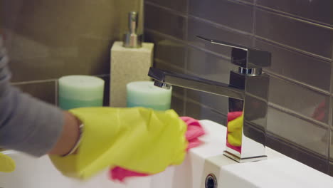 Close-up-of-yellow-gloved-female-hand-wiping-down-faucet-with-pink-microfibre-cloth