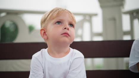 a little boy playfully makes funny eye movements while sitting and raising his hand slightly, with a partial view of someone next to him