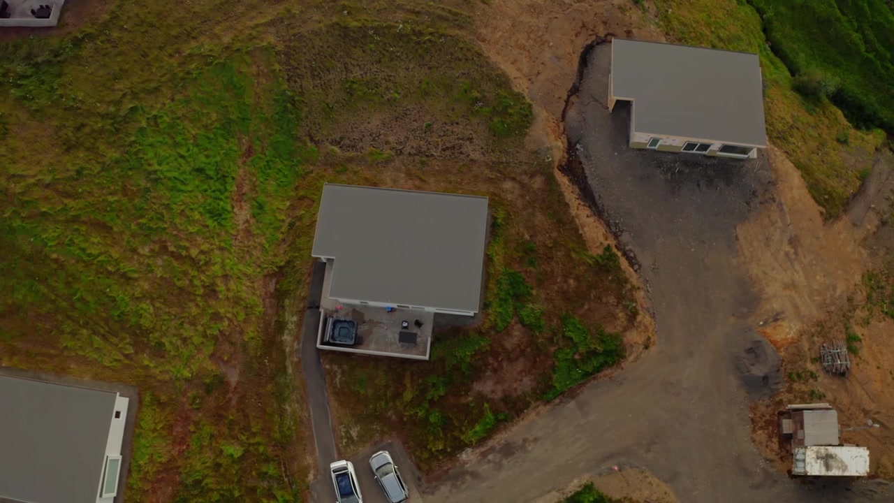 Luftfoto fra drone, der flyver over moderne beboelseshuse bygget på en grøn bakkeskråning nær Akureyri, Island. To biler er parkeret uden for et hus
