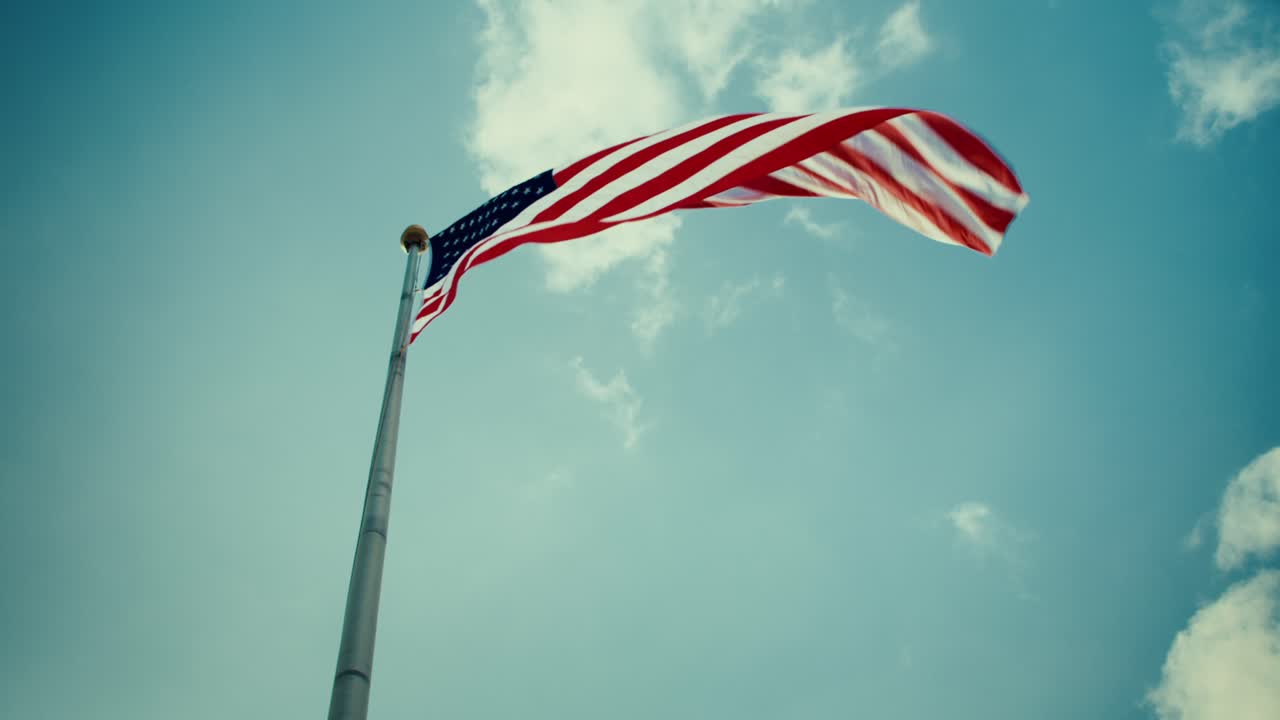 Bandiera americana che sventola su un'alta asta contro un cielo azzurro e nuvole, simbolo patriottico di libertà degli Stati Uniti