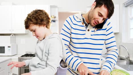 padre hablando en el teléfono móvil mientras corta verduras y hijo usando tableta digital