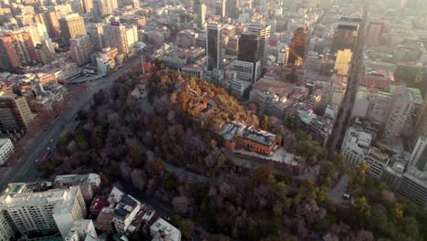 Aerial-orbit-of-Hidalgo-Castle-on-top-of-Santa-Lucia-Hill-covered-in-autumnal-trees-surrounded-by-Santiago-buildings-at-sunset,-Chile