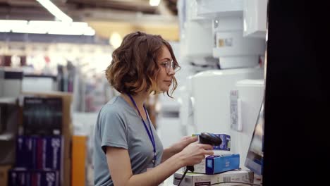 Curly-haired-slender-woman-seller-consultant-in-household-department-scan-by-barcode-stationery-goods-in-the-boxes.-Side-view