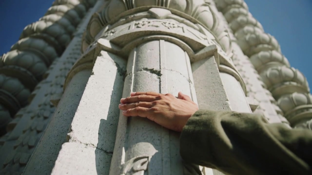 Mano di una persona premuta piatta su una colonna di pietra intagliata su una facciata soleggiata, mentre la telecamera si allontana e si inclina verso l'alto