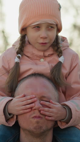 momento sereno como una niña con una gorra rosa cubre tranquilamente los ojos de su padre mientras está sentada en sus hombros en un parque