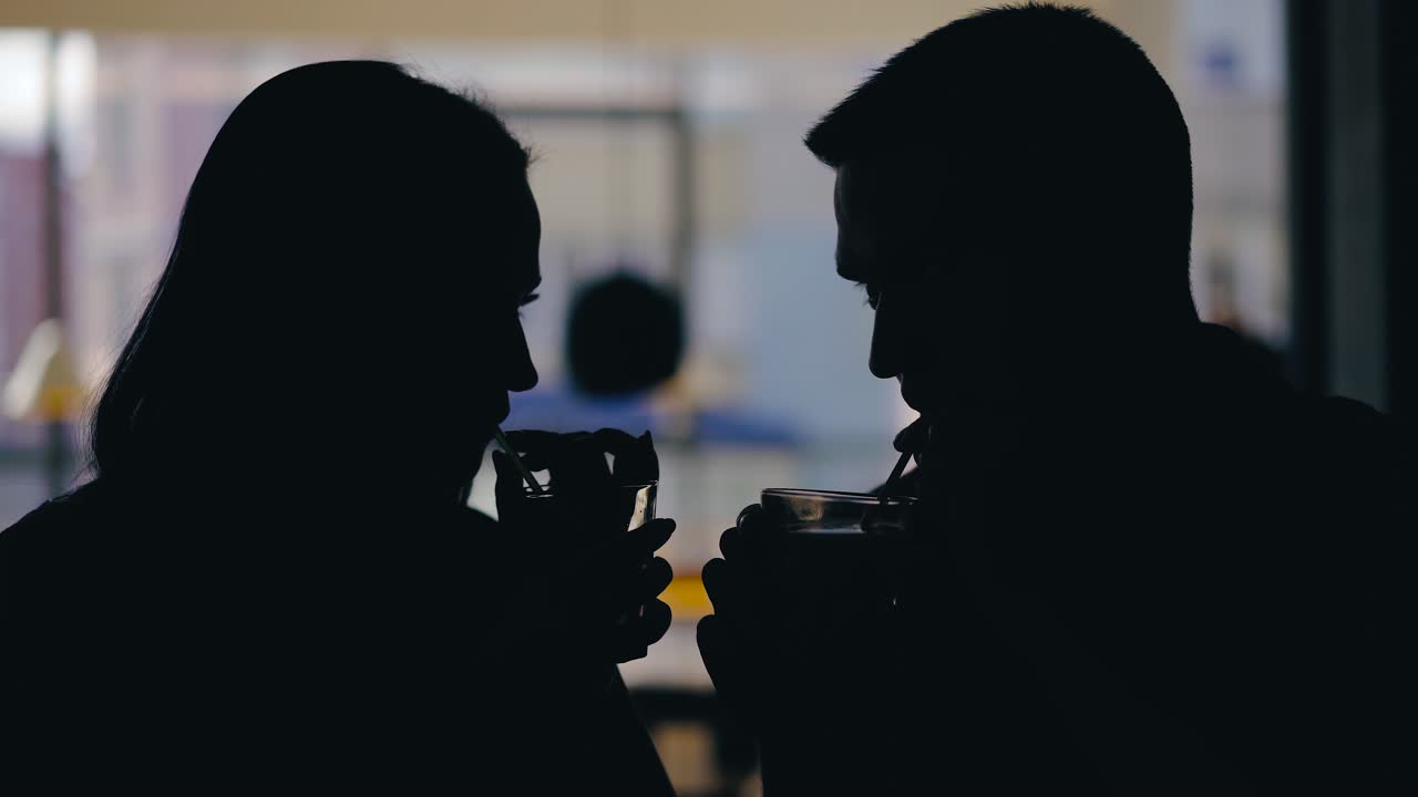 A man and a woman are drinking drinks from tubules on a blurred background in a cafe. Love story. Silhouette of lovers
