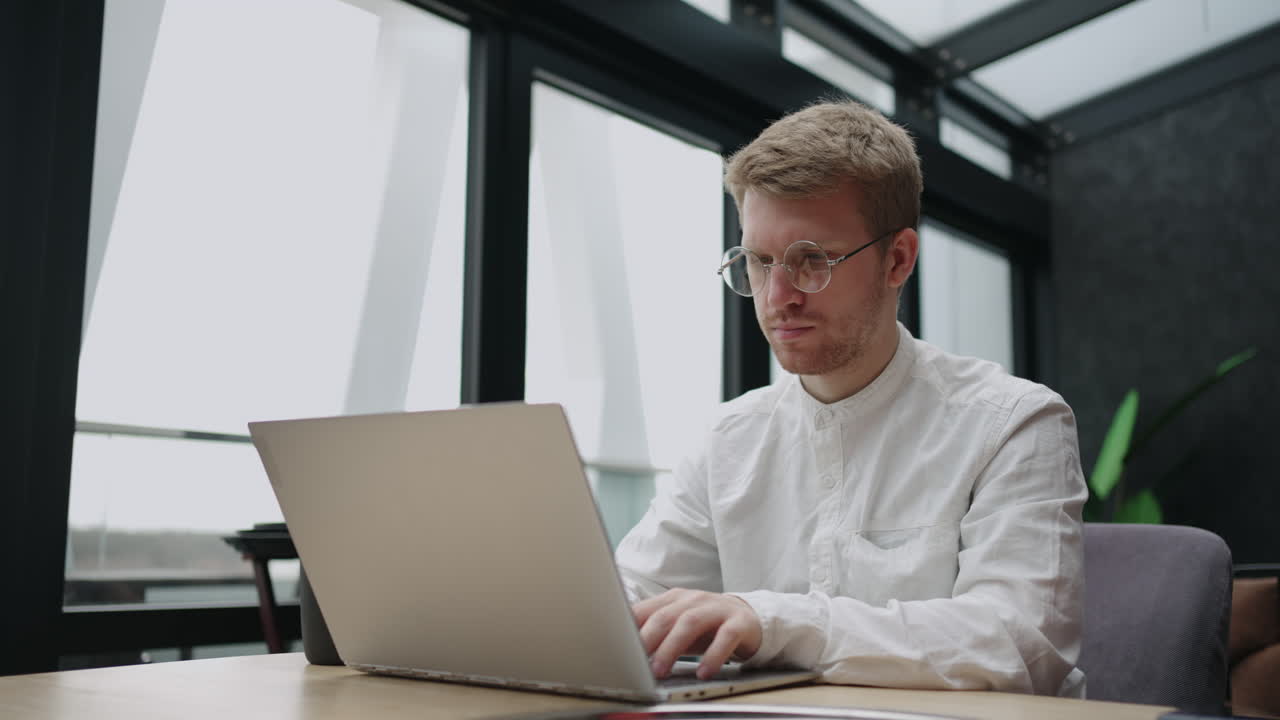 Serious focused determined young bearded businessman or freelancer in glasses and typing on laptop working on project researching writing email to client