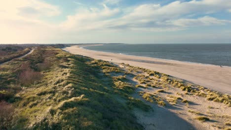 Beautiful-aerial-shot-flying-over-green-dunes-and-a-nearly-empty-beach-at-sunset