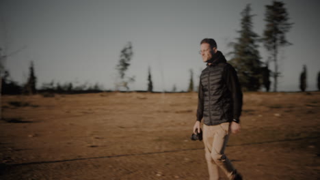 motivated young caucasian male photographer out in nature, holding a camera in his hand and walking toward a forest to take landscape pictures at sunset