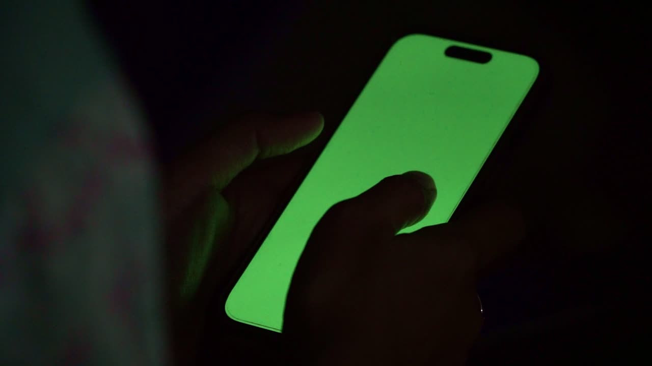 Person using smartphone with green screen display in dark environment, suitable for screen replacement and tech presentations