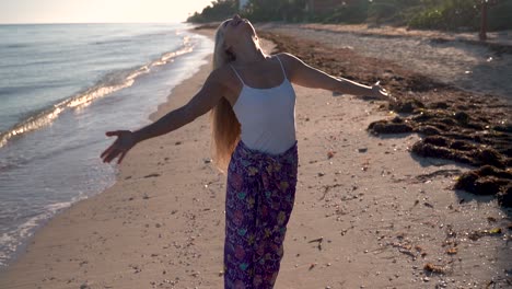 mature blonde woman raises her arms and looks to the sky while on a beach as the camera turns around her