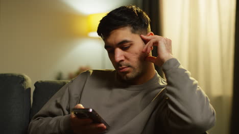 joven que pasa la noche en casa sentado en el sofá con el teléfono móvil desplazándose por internet o medios sociales transmitiendo contenido 4
