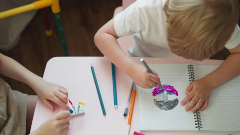 Un-Niño-Pequeño-Dibuja-Mientras-Su-Hermana-Coloca-Etiquetas-En-Un-Borrador-En-El-Escritorio