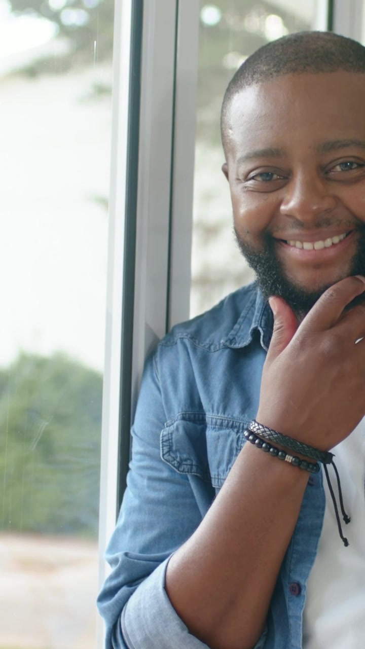 Vertical video: Camera filming man beside window in denim rolling sleeves, touching chin, smiling