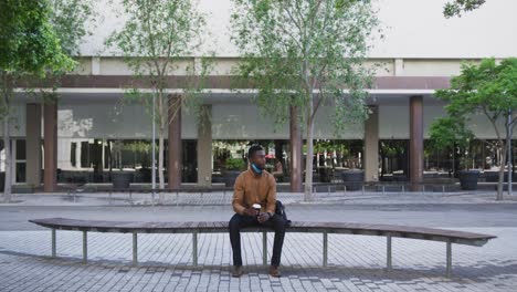 african american businessman with face mask holding coffee sitting on bench