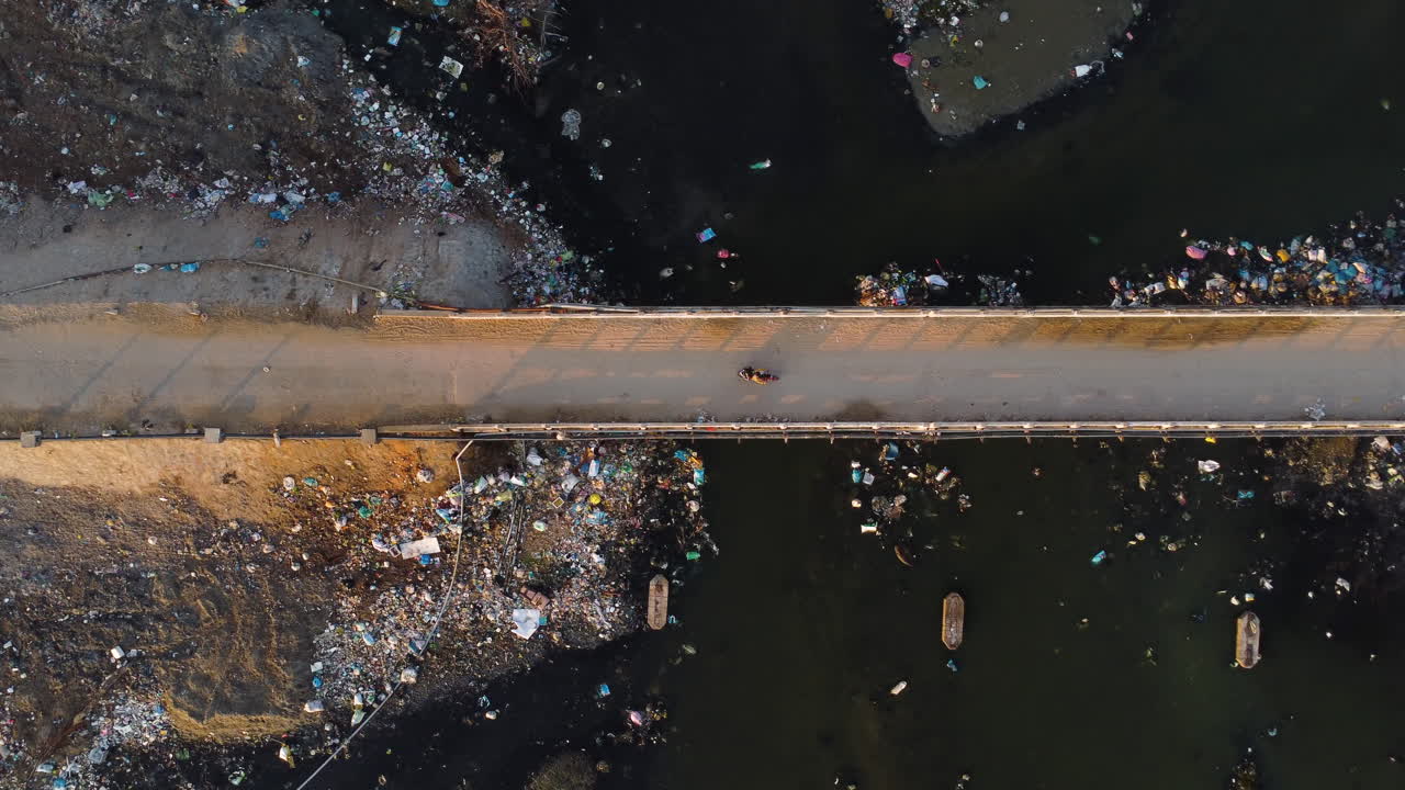 Aerial top down of old narrowed suspension bridge with scooter driving over a polluted river filled with garbage plastic waste pollution trash