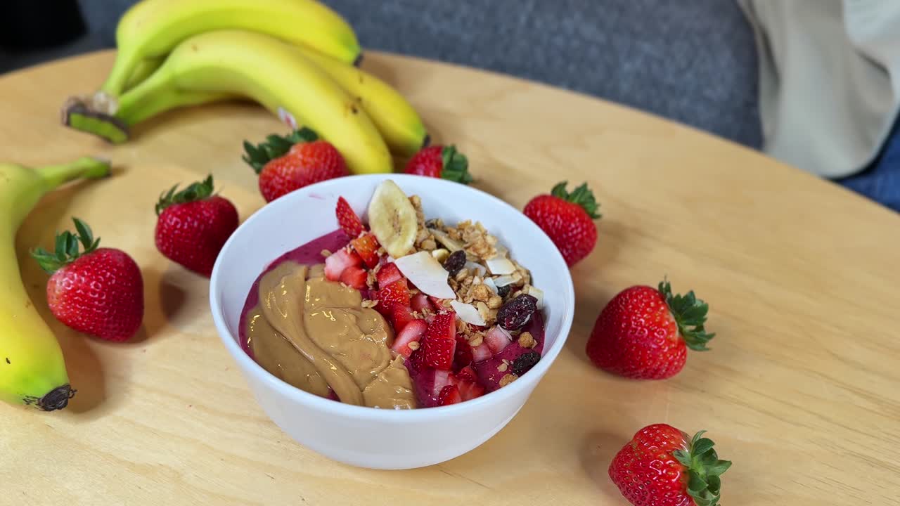 Una mano con un guanto aggiunge una banana a una densa ciotola di açaí con burro di arachidi, fragole, granola e cocco: un primo piano pulito e appetitoso, ideale per video di ricette, promozioni di bar e contenuti sul benessere