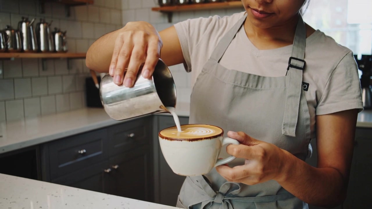 Barista con grembiule grigio che inclina una brocca e versa il latte al bancone del caffè, creando latte art nella tazza.