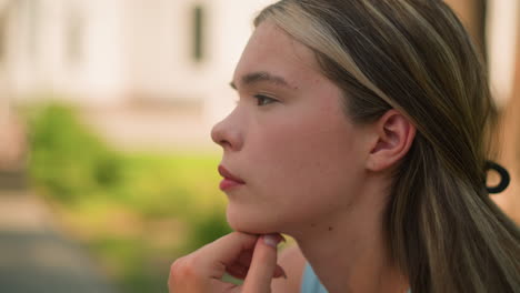 side view of young woman with hand under chin, looking thoughtful while glancing into distance, with blurred greenery background and building