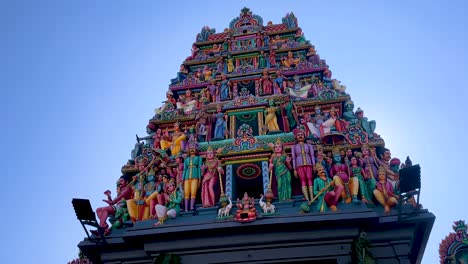 Camera tilts upward to reveal a vibrant, intricately decorated Hindu temple gopuram under bright daylight, highlighting detailed sculptures and urban surroundings