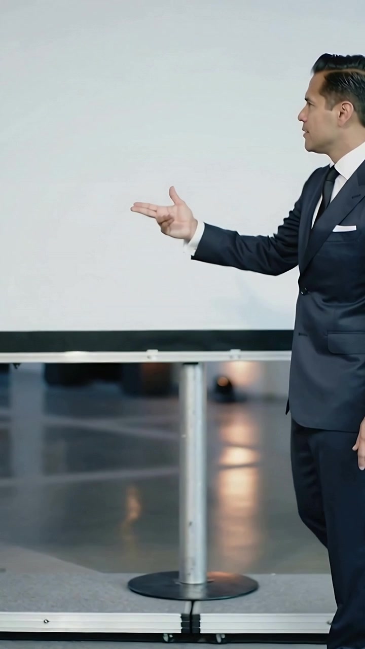 Video verticale: Presentatore che cammina in abito blu navy, indicando attraverso il palco per evidenziare lo schermo.