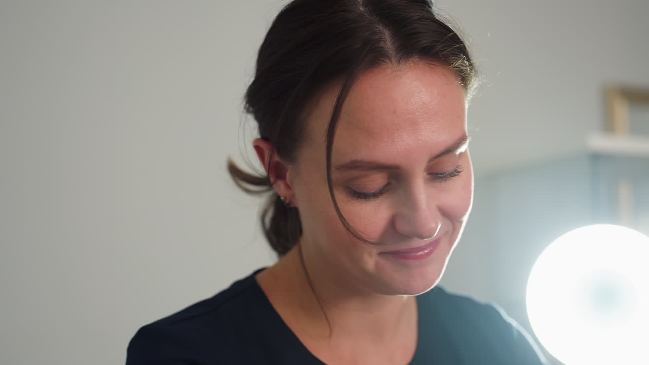 Portrait joyful beauty technician wearing navy blue work dress standing in spa room under bright backlight focused while working showing professionalism through warm smile and concentrated gaze