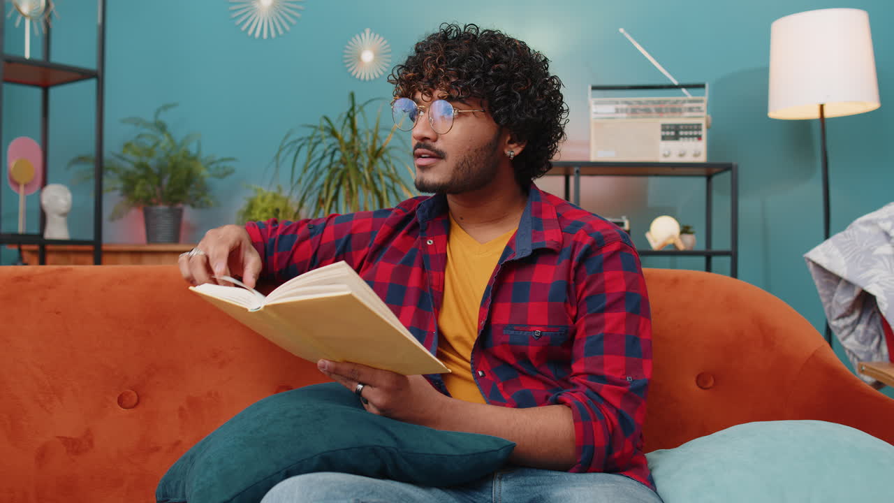 Indian man reading interesting book turning pages smiling enjoying literature taking a rest on sofa