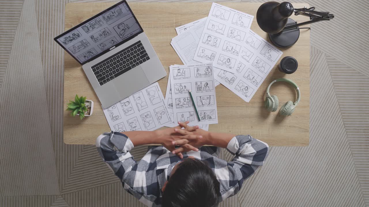 vista superior de un artista masculino asiático estirándose mientras dibuja un guión gráfico para la película en la mesa con una computadora portátil y auriculares en el estudio