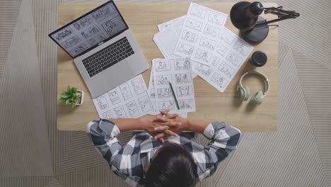 top view of asian male artist stretching while drawing storyboard for the film on the table with a laptop and headphones in the studio