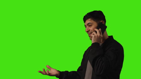 smiling and animated young man talking on mobile phone walking into frame against green screen studio background