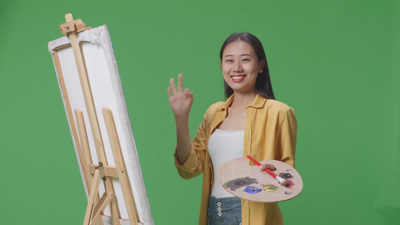 아시아 여성 예술가가 미소 짓고 녹색 스크린 스튜디오에서 오일 페인트와 브러쉬로 캔버스에 그림을 그리는 동안 카메라에 은 몸짓을 보여줍니다.