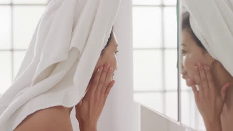 video of portrait of smiling biracial woman with towel on hair applying cream in bathroom