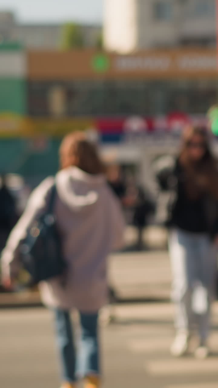 Donna bianca con felpa con cappuccio che cammina su un attraversamento pedonale urbano trafficato, scatto sincero da dietro, luce solare mattutina, folla sfocata, borsa a tracolla, jeans, sneakers, ritmo al rallentatore, tragitto giornaliero, stato d'animo riflessivo