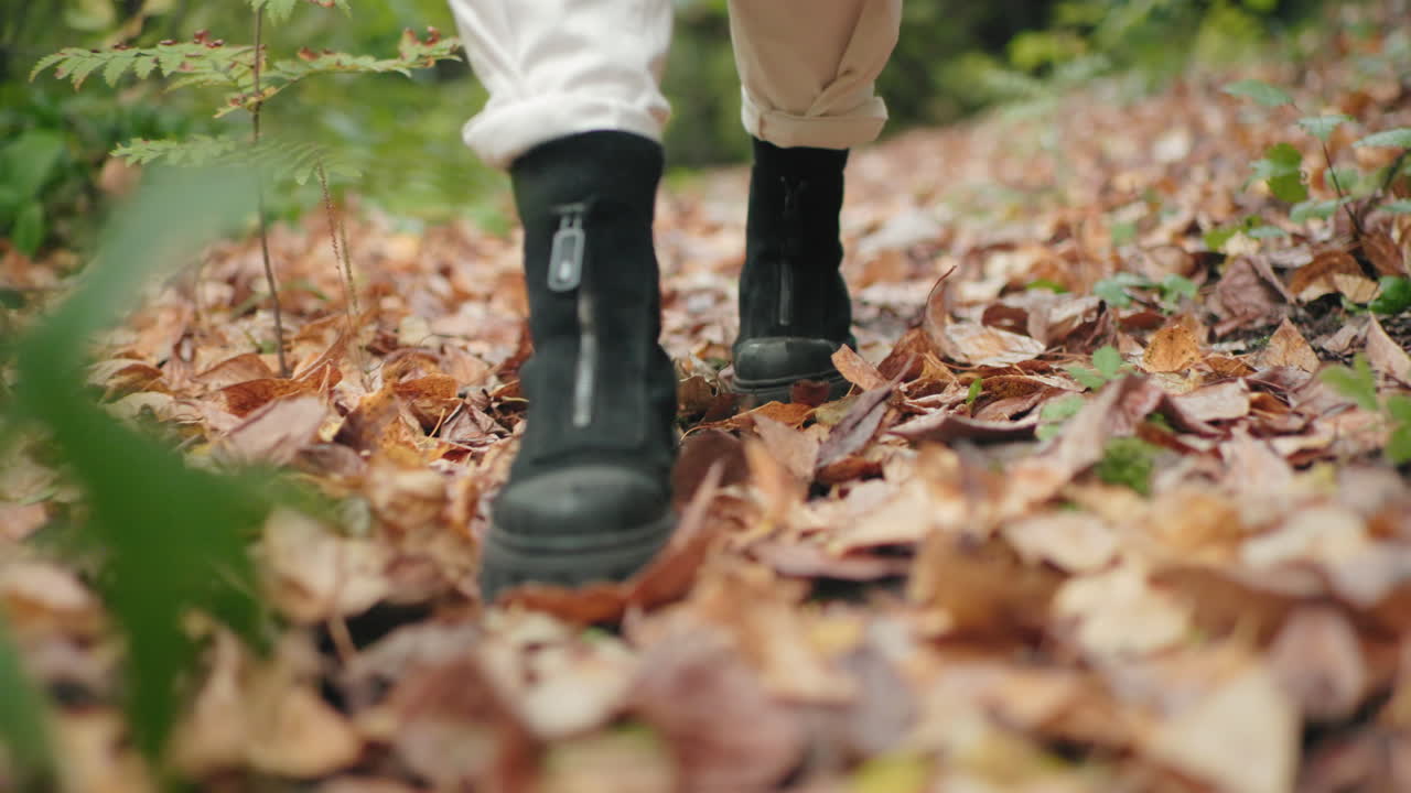 Vista de las piernas de un visitante del bosque con botas negras paseando por un sendero forestal sobre hojas secas; la perspectiva cercana al suelo muestra los pasos y el movimiento, ambiente otoñal acogedor, bokeh suave, paseo tranquilo por la naturaleza, ambiente de mindfulness