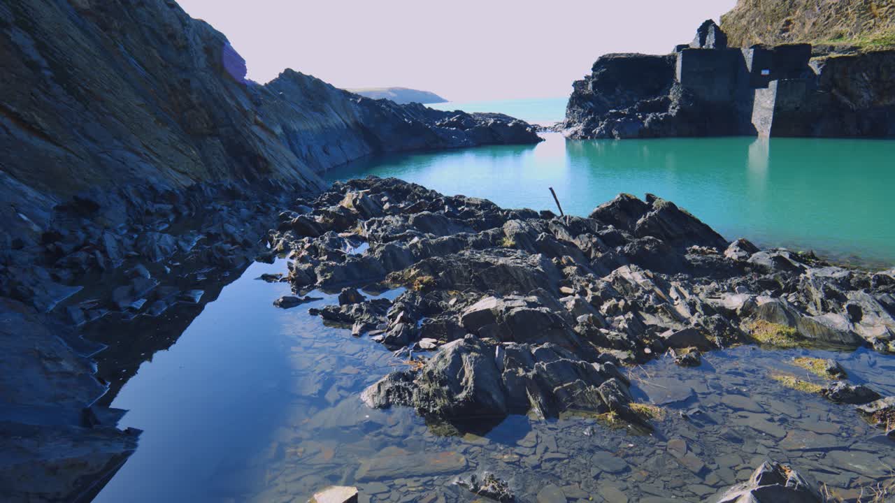 Panning Up Across Seaside Historic Blue Lagoon Quarry with Calm Water and Surrounding Rocks with Old Buildings and Cliffs with Sea in Background.